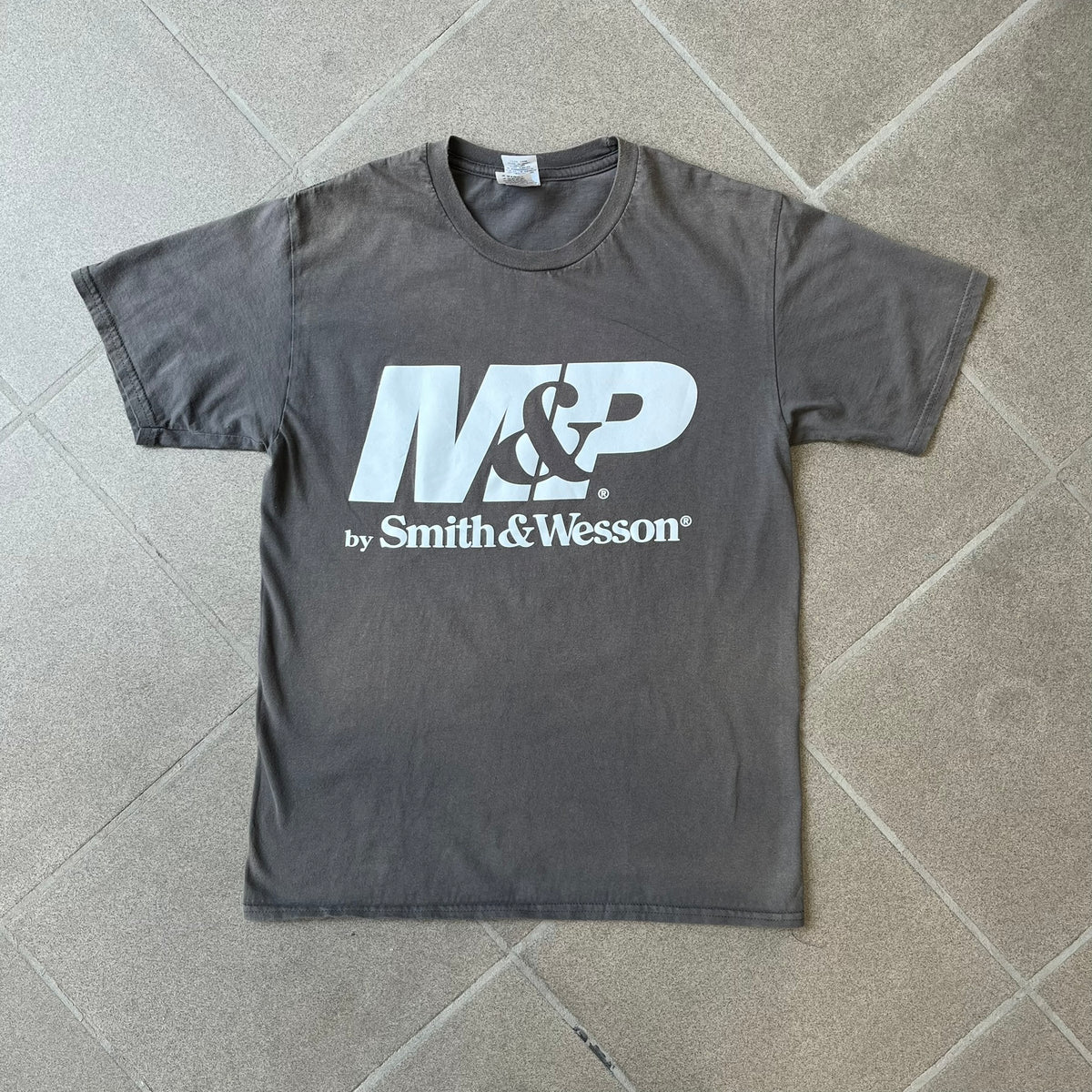 M&P by Smith & Wesson Gray Tee features a washed grey finish with a large M&P logo printed across the front and smaller "by Smith & Wesson" text underneath. Made from 100% cotton on a Gildan blank