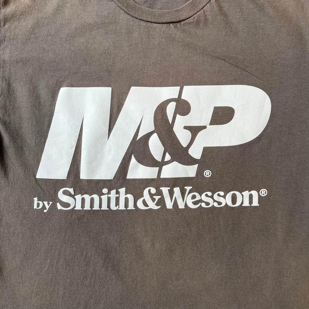 M&P by Smith & Wesson Gray Tee features a washed grey finish with a large M&P logo printed across the front and smaller "by Smith & Wesson" text underneath. Made from 100% cotton on a Gildan blank