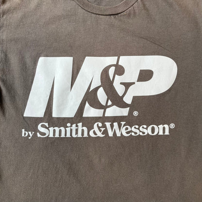 M&P by Smith & Wesson Gray Tee features a washed grey finish with a large M&P logo printed across the front and smaller "by Smith & Wesson" text underneath. Made from 100% cotton on a Gildan blank