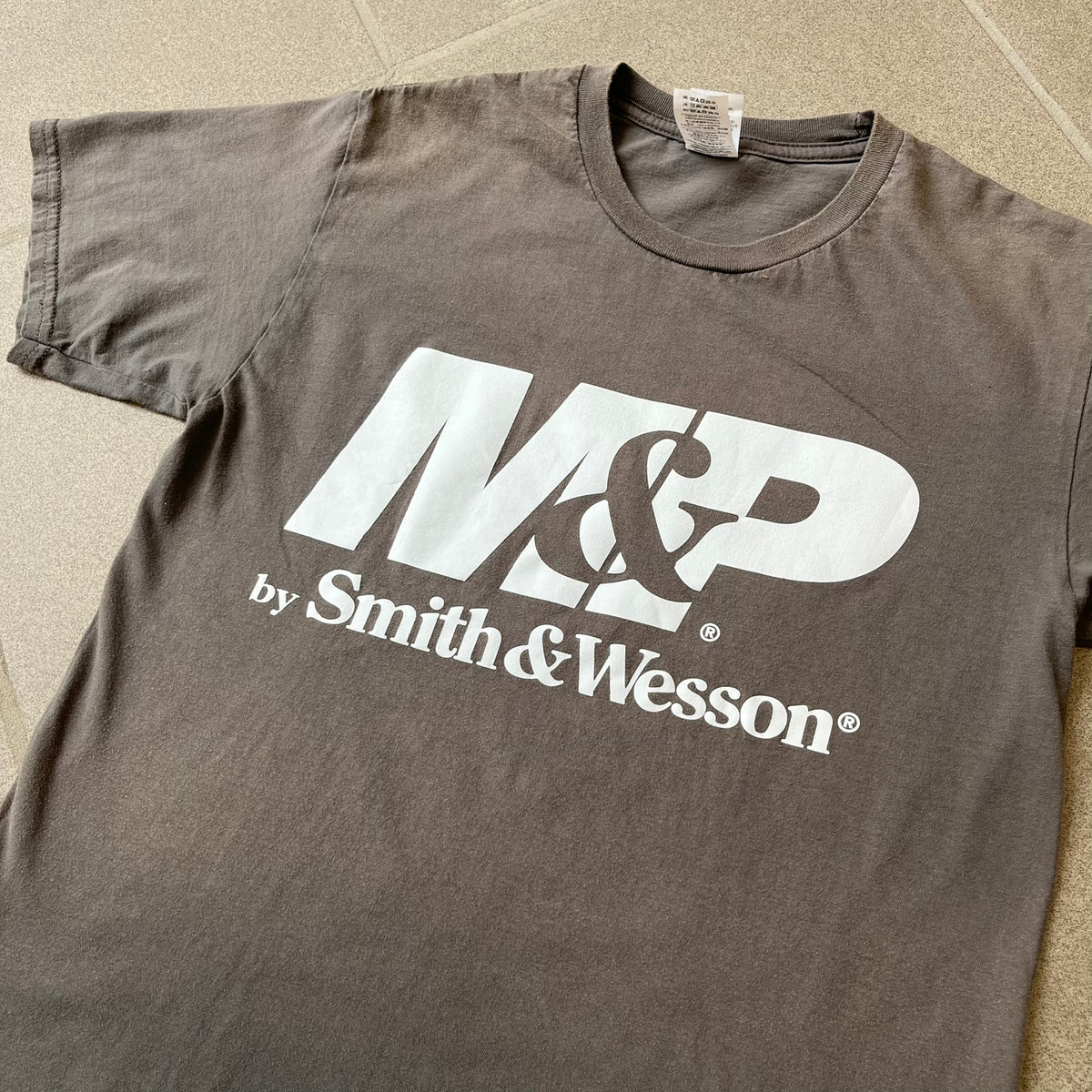 M&P by Smith & Wesson Gray Tee features a washed grey finish with a large M&P logo printed across the front and smaller "by Smith & Wesson" text underneath. Made from 100% cotton on a Gildan blank