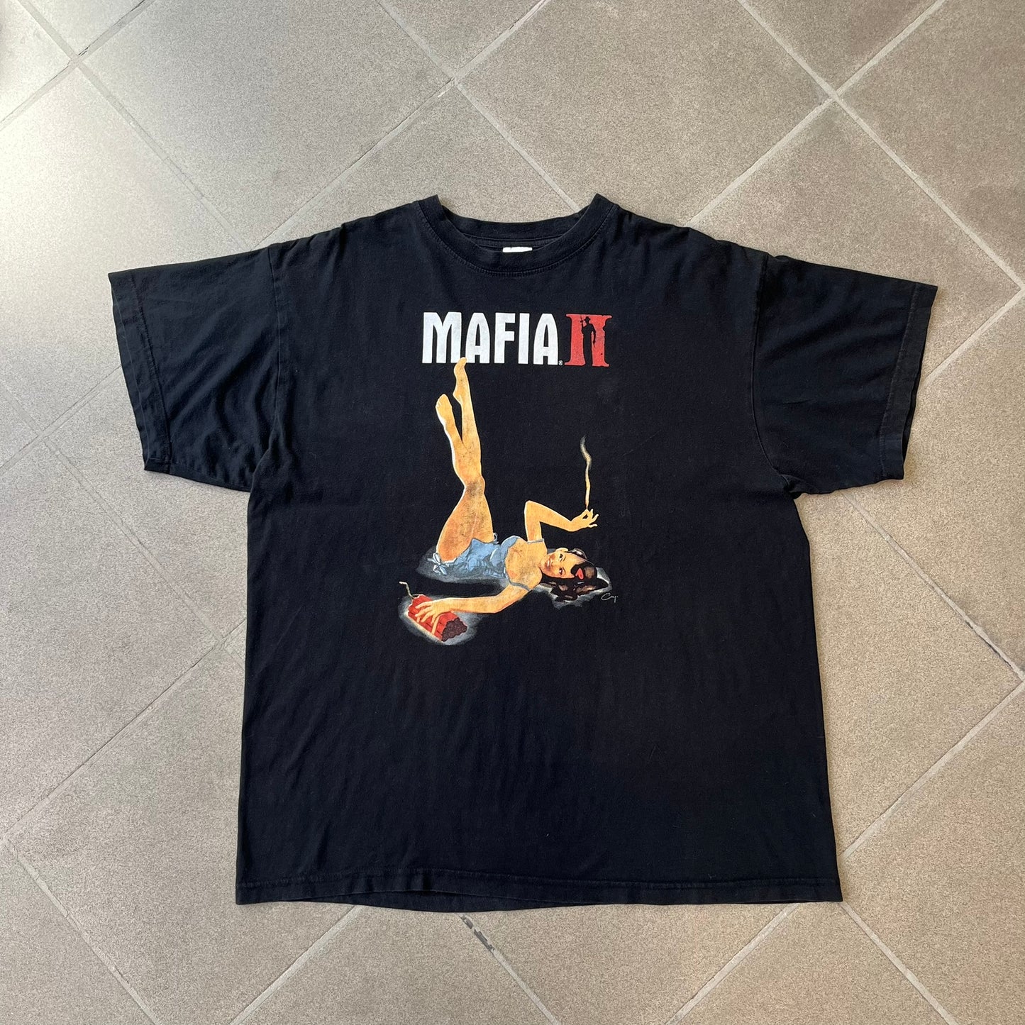 Mafia 2 Smoking Woman 2010 Black Promo Tee features a black T-shirt made on a Stedman blank. The front has a large graphic of a woman lying down while smoking a cigarette and holding dynamite in the other hand. A white and red Mafia II logo is printed above the graphic.
