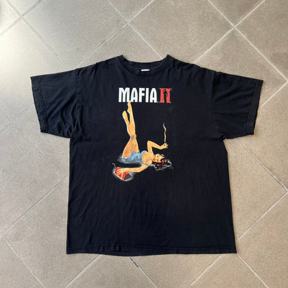 Mafia 2 Smoking Woman 2010 Black Promo Tee features a black T-shirt made on a Stedman blank. The front has a large graphic of a woman lying down while smoking a cigarette and holding dynamite in the other hand. A white and red Mafia II logo is printed above the graphic.
