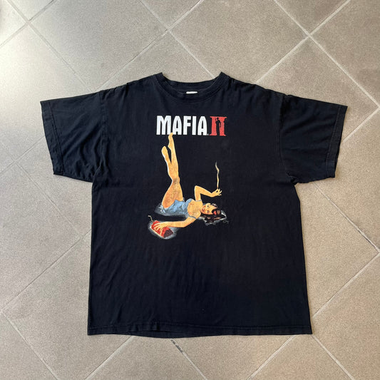 Mafia 2 Smoking Woman 2010 Black Promo Tee features a black T-shirt made on a Stedman blank. The front has a large graphic of a woman lying down while smoking a cigarette and holding dynamite in the other hand. A white and red Mafia II logo is printed above the graphic.
