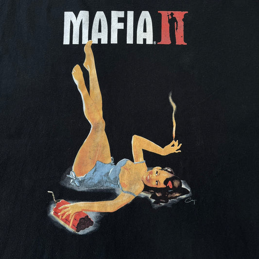 Mafia 2 Smoking Woman 2010 Black Promo Tee features a black T-shirt made on a Stedman blank. The front has a large graphic of a woman lying down while smoking a cigarette and holding dynamite in the other hand. A white and red Mafia II logo is printed above the graphic.

