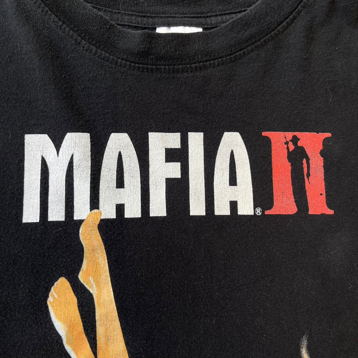 Mafia 2 Smoking Woman 2010 Black Promo Tee features a black T-shirt made on a Stedman blank. The front has a large graphic of a woman lying down while smoking a cigarette and holding dynamite in the other hand. A white and red Mafia II logo is printed above the graphic.
