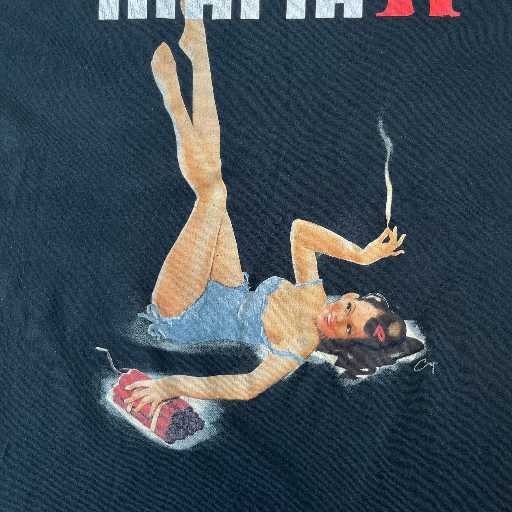 Mafia 2 Smoking Woman 2010 Black Promo Tee features a black T-shirt made on a SG blank. The front has a large graphic of a woman lying down while smoking a cigarette and holding dynamite in the other hand. A white and red Mafia II logo is printed above the graphic.
