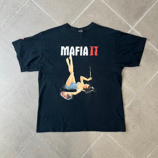 Mafia 2 Smoking Woman 2010 Black Promo Tee features a black T-shirt made on a SG blank. The front has a large graphic of a woman lying down while smoking a cigarette and holding dynamite in the other hand. A white and red Mafia II logo is printed above the graphic.
