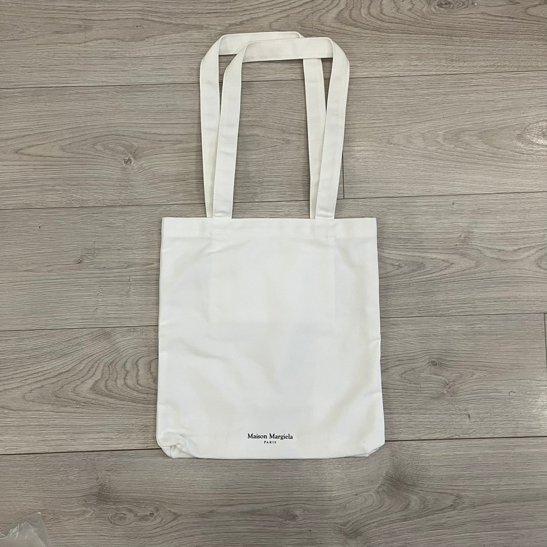 Maison Margiela Replica Tote Bag in off-white with a textured finish inspired by natural jute. Features “Replica” fragrances patch on one side, an interior slip pocket, and small “Maison Margiela Paris” text on the other side. Made in Paris, versatile for daily errands, work, or casual outings.