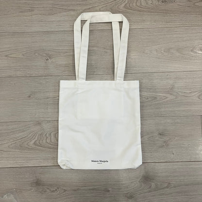 Maison Margiela Replica Tote Bag in off-white with a textured finish inspired by natural jute. Features “Replica” fragrances patch on one side, an interior slip pocket, and small “Maison Margiela Paris” text on the other side. Made in Paris, versatile for daily errands, work, or casual outings.