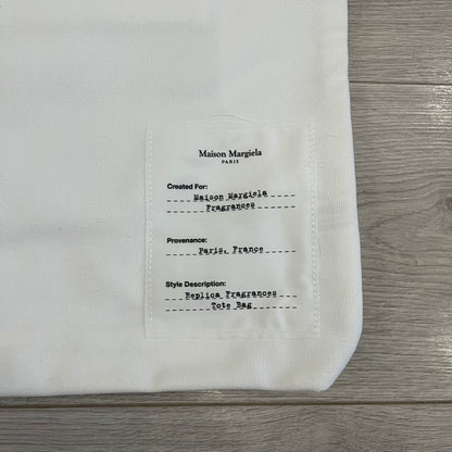 Maison Margiela Replica Tote Bag in off-white with a textured finish inspired by natural jute. Features “Replica” fragrances patch on one side, an interior slip pocket, and small “Maison Margiela Paris” text on the other side. Made in Paris, versatile for daily errands, work, or casual outings.