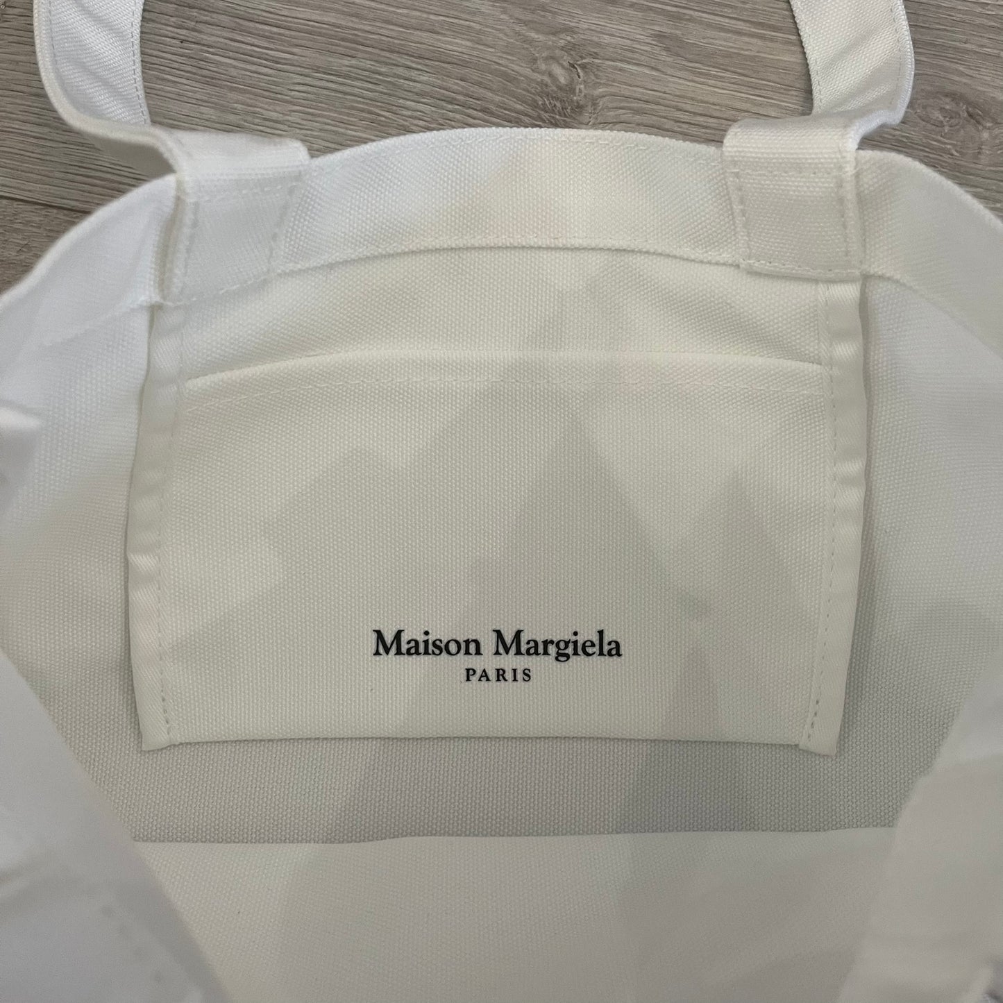 Maison Margiela Replica Tote Bag in off-white with a textured finish inspired by natural jute. Features “Replica” fragrances patch on one side, an interior slip pocket, and small “Maison Margiela Paris” text on the other side. Made in Paris, versatile for daily errands, work, or casual outings.