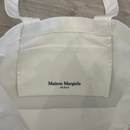 Maison Margiela Replica Tote Bag in off-white with a textured finish inspired by natural jute. Features “Replica” fragrances patch on one side, an interior slip pocket, and small “Maison Margiela Paris” text on the other side. Made in Paris, versatile for daily errands, work, or casual outings.