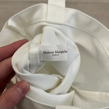 Maison Margiela Replica Tote Bag in off-white with a textured finish inspired by natural jute. Features “Replica” fragrances patch on one side, an interior slip pocket, and small “Maison Margiela Paris” text on the other side. Made in Paris, versatile for daily errands, work, or casual outings.