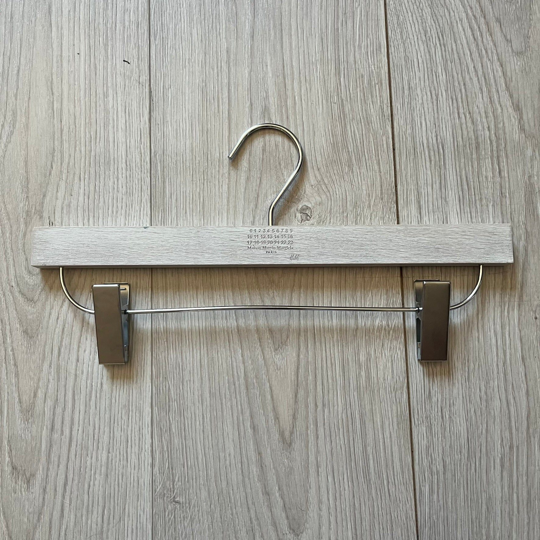 Maison Margiela x H&amp;M 2012 hanger in a white-washed wood finish with engraved logos. Features a straight wooden bar with metal clips, perfect for hanging pants, skirts, or shorts. A rare, functional collectible from this iconic collaboration.