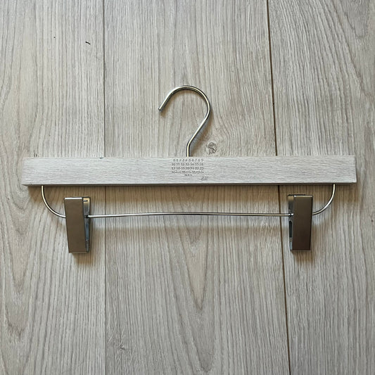 Maison Margiela x H&amp;M 2012 hanger in a white-washed wood finish with engraved logos. Features a straight wooden bar with metal clips, perfect for hanging pants, skirts, or shorts. A rare, functional collectible from this iconic collaboration.