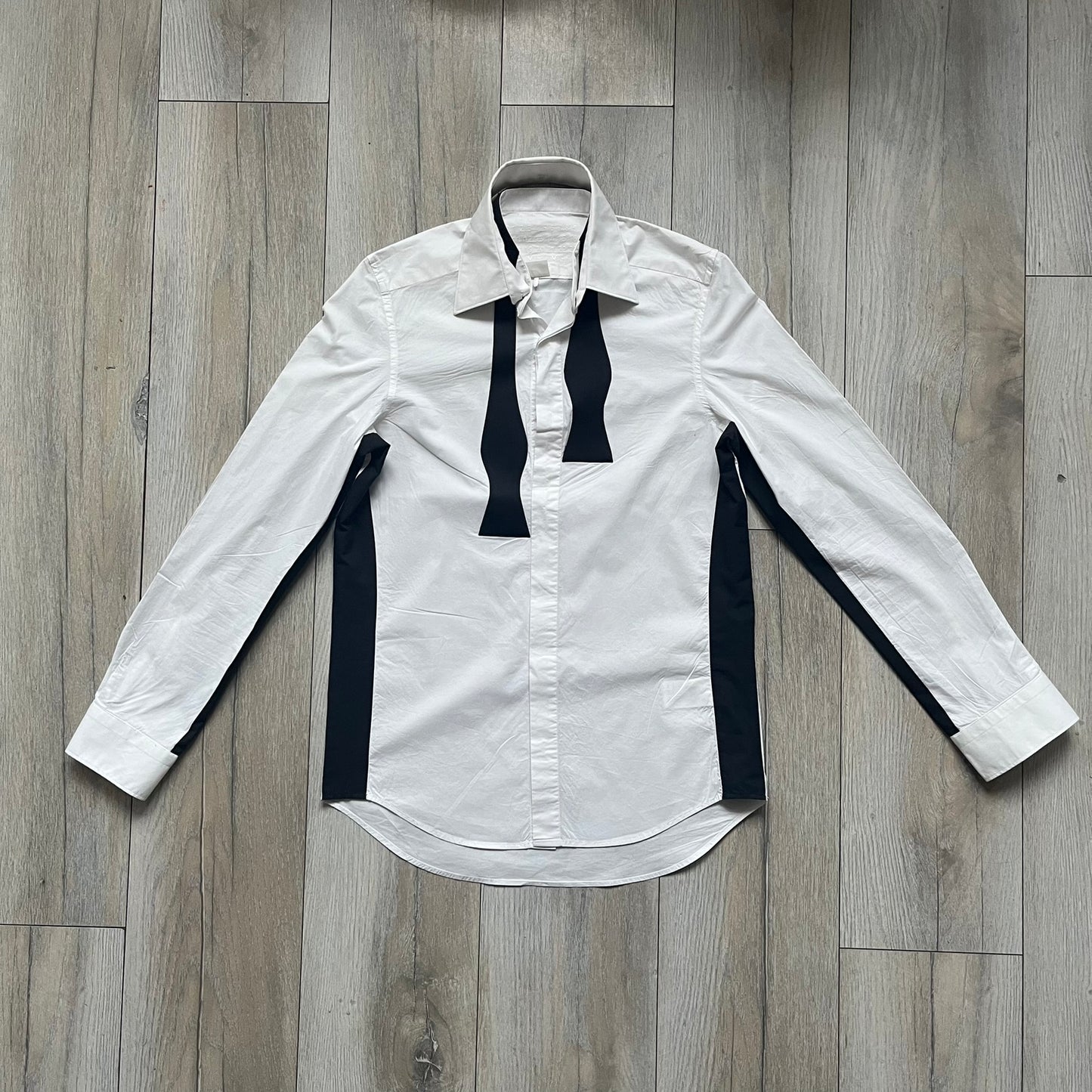 White long-sleeve shirt from the Maison Margiela x H&amp;M Autumn/Winter 2012 collaboration, constructed from 100% cotton. The design features a permanently sewn-in black bow tie positioned loosely at the collar, creating a trompe l’oeil effect that gives the appearance of a casually undone formal look. Additional details include black side panels and underarm stripes for contrast, as well as a detachable collar.