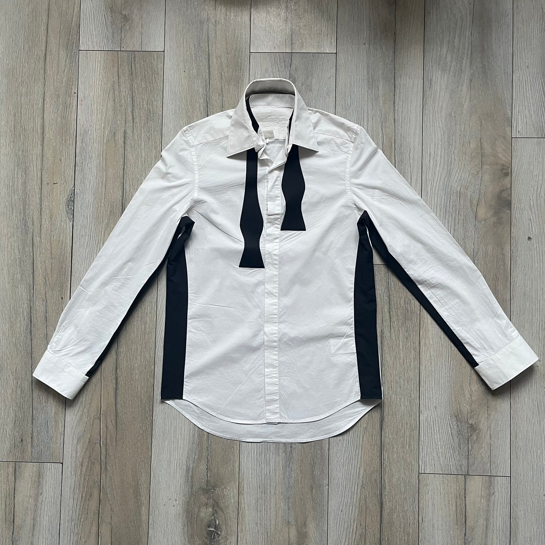 White long-sleeve shirt from the Maison Margiela x H&amp;M Autumn/Winter 2012 collaboration, constructed from 100% cotton. The design features a permanently sewn-in black bow tie positioned loosely at the collar, creating a trompe l’oeil effect that gives the appearance of a casually undone formal look. Additional details include black side panels and underarm stripes for contrast, as well as a detachable collar.