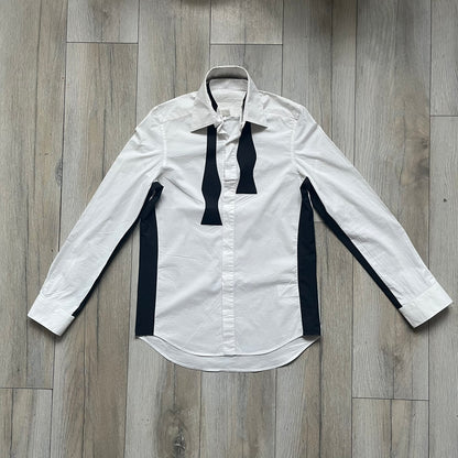 White long-sleeve shirt from the Maison Margiela x H&amp;M Autumn/Winter 2012 collaboration, constructed from 100% cotton. The design features a permanently sewn-in black bow tie positioned loosely at the collar, creating a trompe l’oeil effect that gives the appearance of a casually undone formal look. Additional details include black side panels and underarm stripes for contrast, as well as a detachable collar.