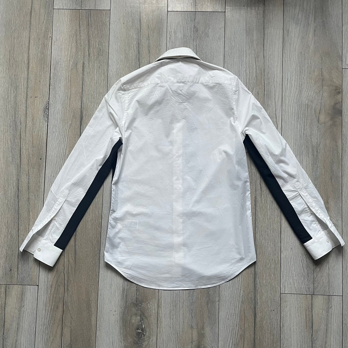 White long-sleeve shirt from the Maison Margiela x H&amp;M Autumn/Winter 2012 collaboration, constructed from 100% cotton. The design features a permanently sewn-in black bow tie positioned loosely at the collar, creating a trompe l’oeil effect that gives the appearance of a casually undone formal look. Additional details include black side panels and underarm stripes for contrast, as well as a detachable collar.