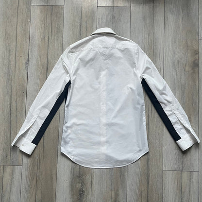 White long-sleeve shirt from the Maison Margiela x H&amp;M Autumn/Winter 2012 collaboration, constructed from 100% cotton. The design features a permanently sewn-in black bow tie positioned loosely at the collar, creating a trompe l’oeil effect that gives the appearance of a casually undone formal look. Additional details include black side panels and underarm stripes for contrast, as well as a detachable collar.