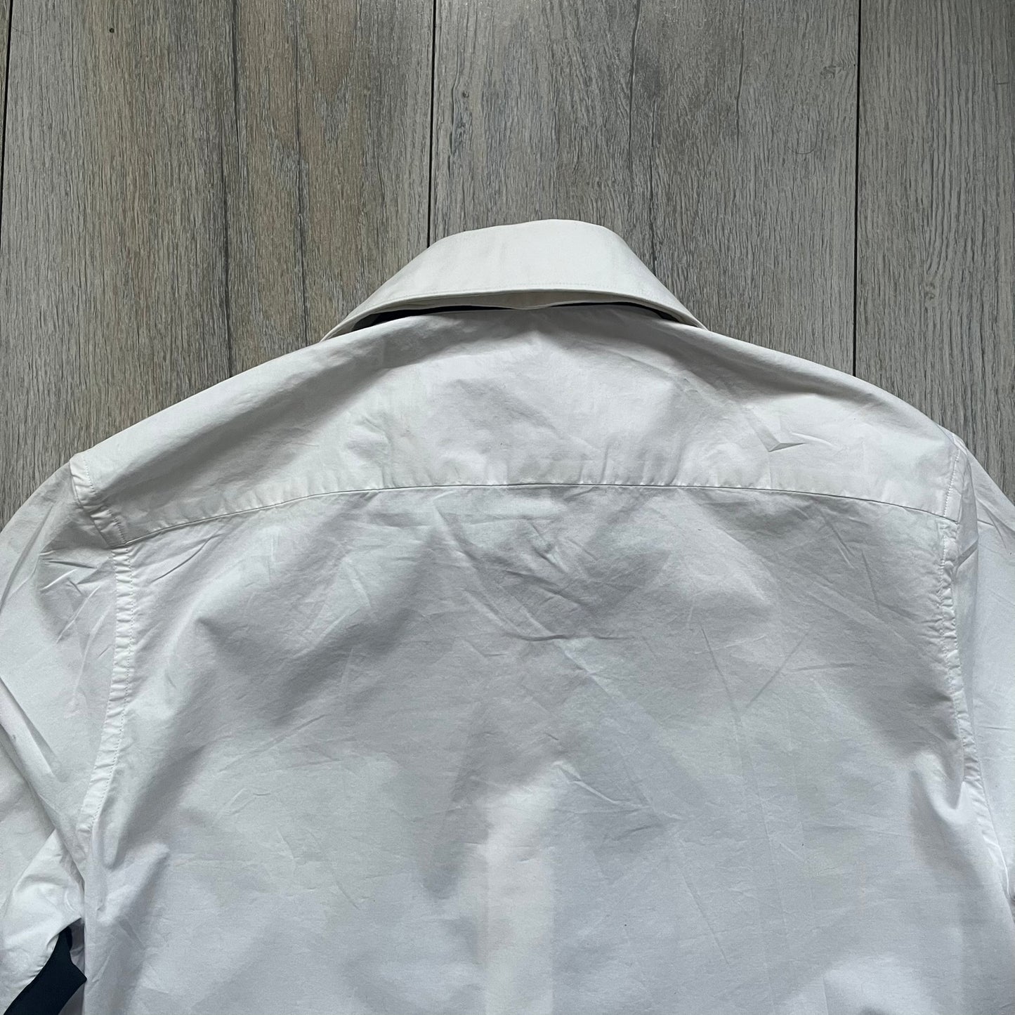 White long-sleeve shirt from the Maison Margiela x H&amp;M Autumn/Winter 2012 collaboration, constructed from 100% cotton. The design features a permanently sewn-in black bow tie positioned loosely at the collar, creating a trompe l’oeil effect that gives the appearance of a casually undone formal look. Additional details include black side panels and underarm stripes for contrast, as well as a detachable collar.