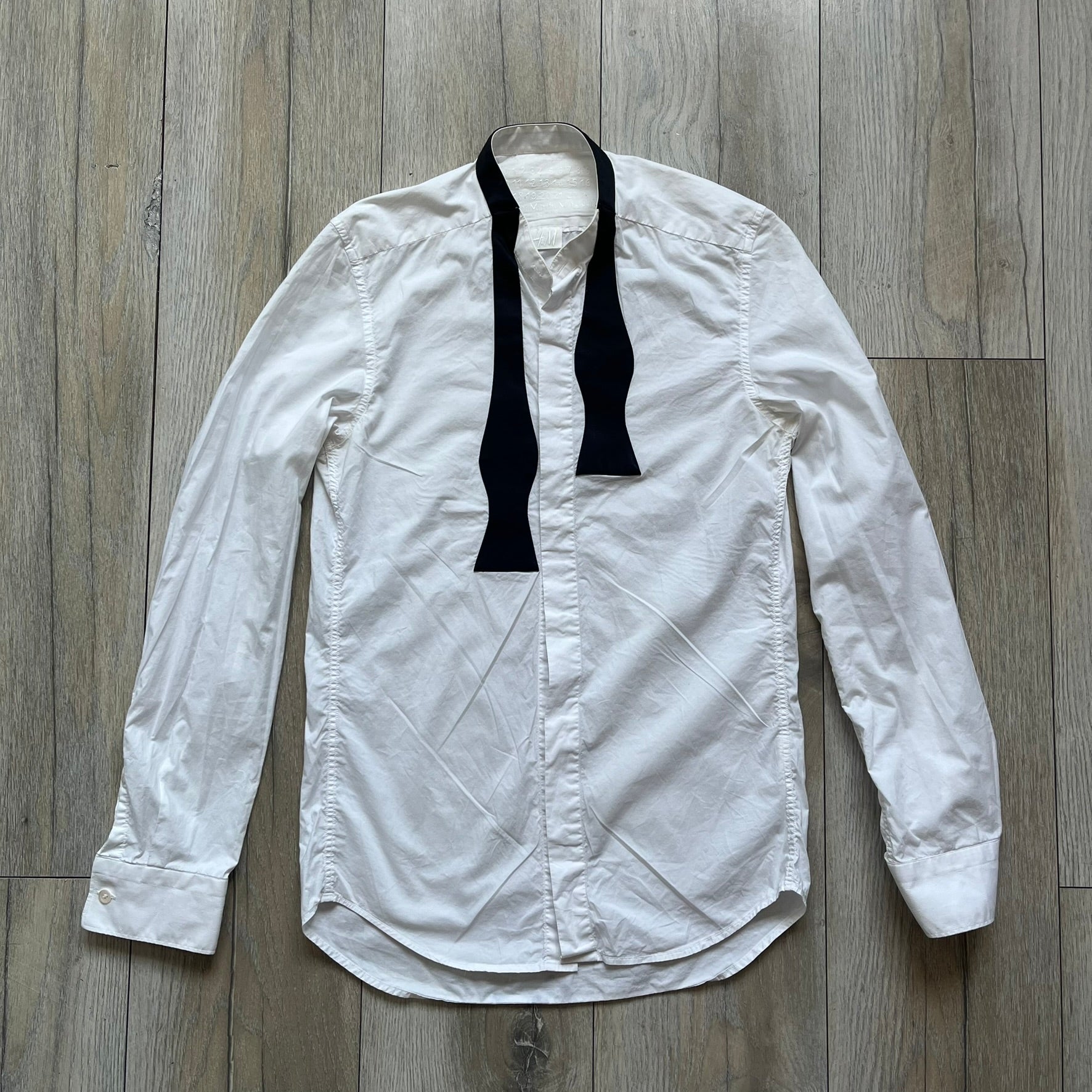 White long-sleeve shirt from the Maison Margiela x H&amp;M Autumn/Winter 2012 collaboration, constructed from 100% cotton. The design features a permanently sewn-in black bow tie positioned loosely at the collar, creating a trompe l’oeil effect that gives the appearance of a casually undone formal look.&nbsp;