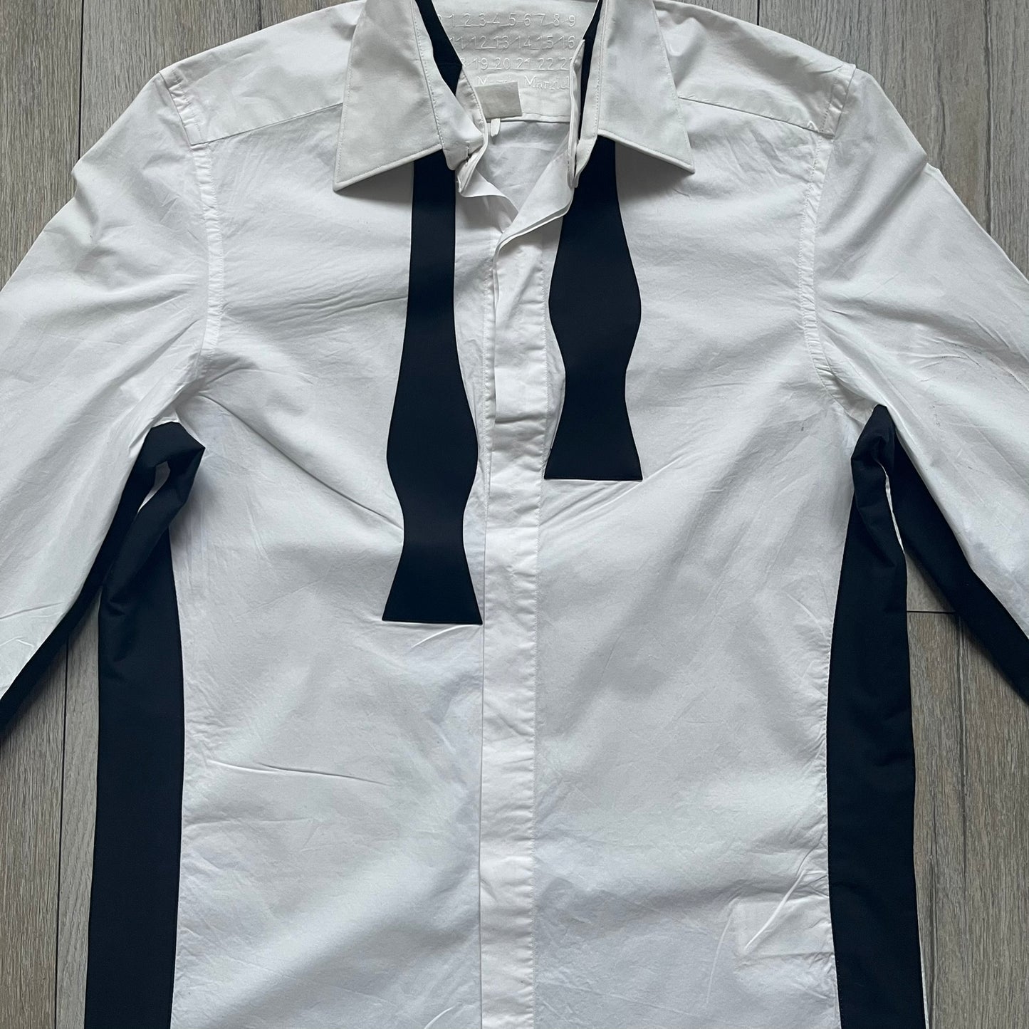 White long-sleeve shirt from the Maison Margiela x H&amp;M Autumn/Winter 2012 collaboration, constructed from 100% cotton. The design features a permanently sewn-in black bow tie positioned loosely at the collar, creating a trompe l’oeil effect that gives the appearance of a casually undone formal look. Additional details include black side panels and underarm stripes for contrast, as well as a detachable collar.