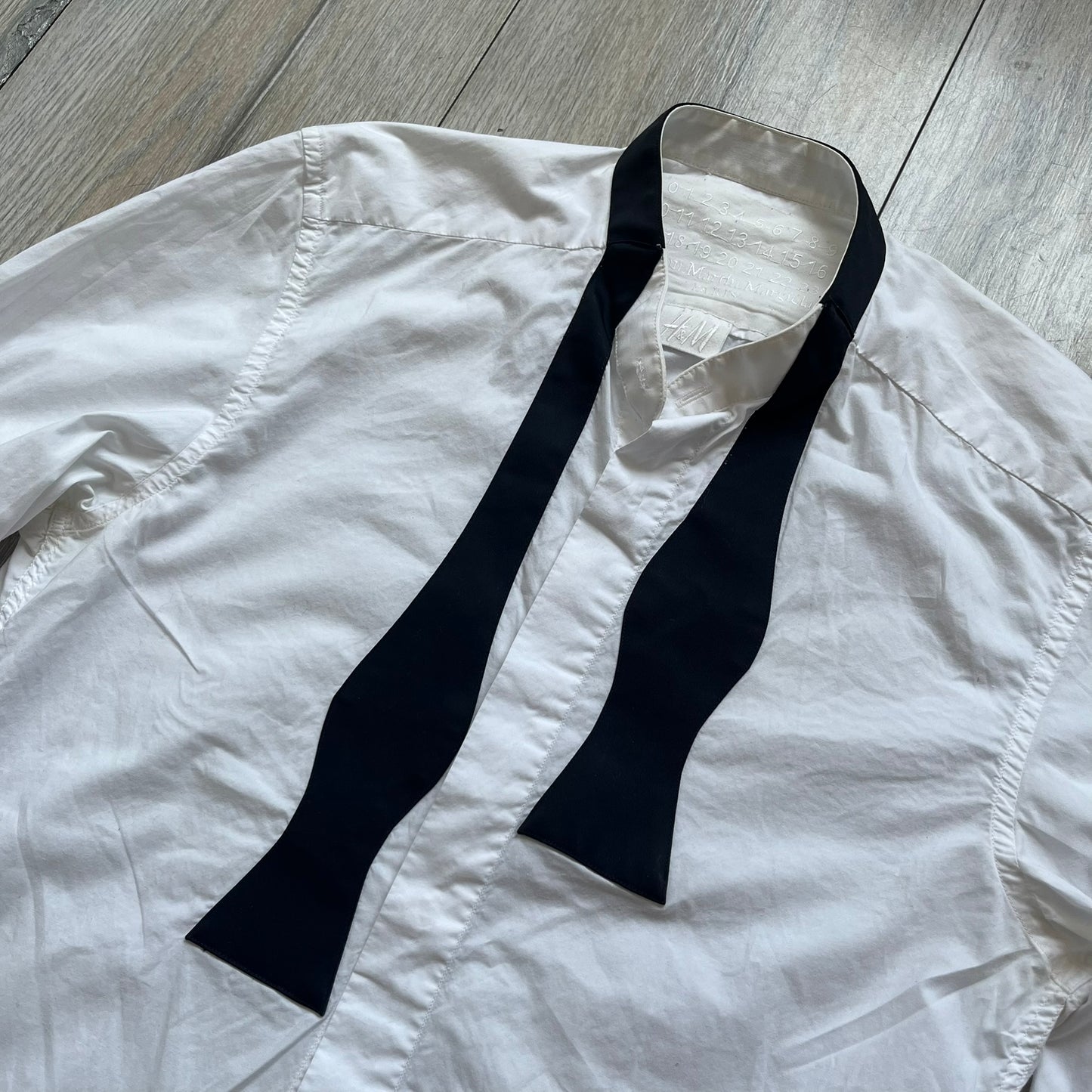 White long-sleeve shirt from the Maison Margiela x H&amp;M Autumn/Winter 2012 collaboration, constructed from 100% cotton. The design features a permanently sewn-in black bow tie positioned loosely at the collar, creating a trompe l’oeil effect that gives the appearance of a casually undone formal look.&nbsp;