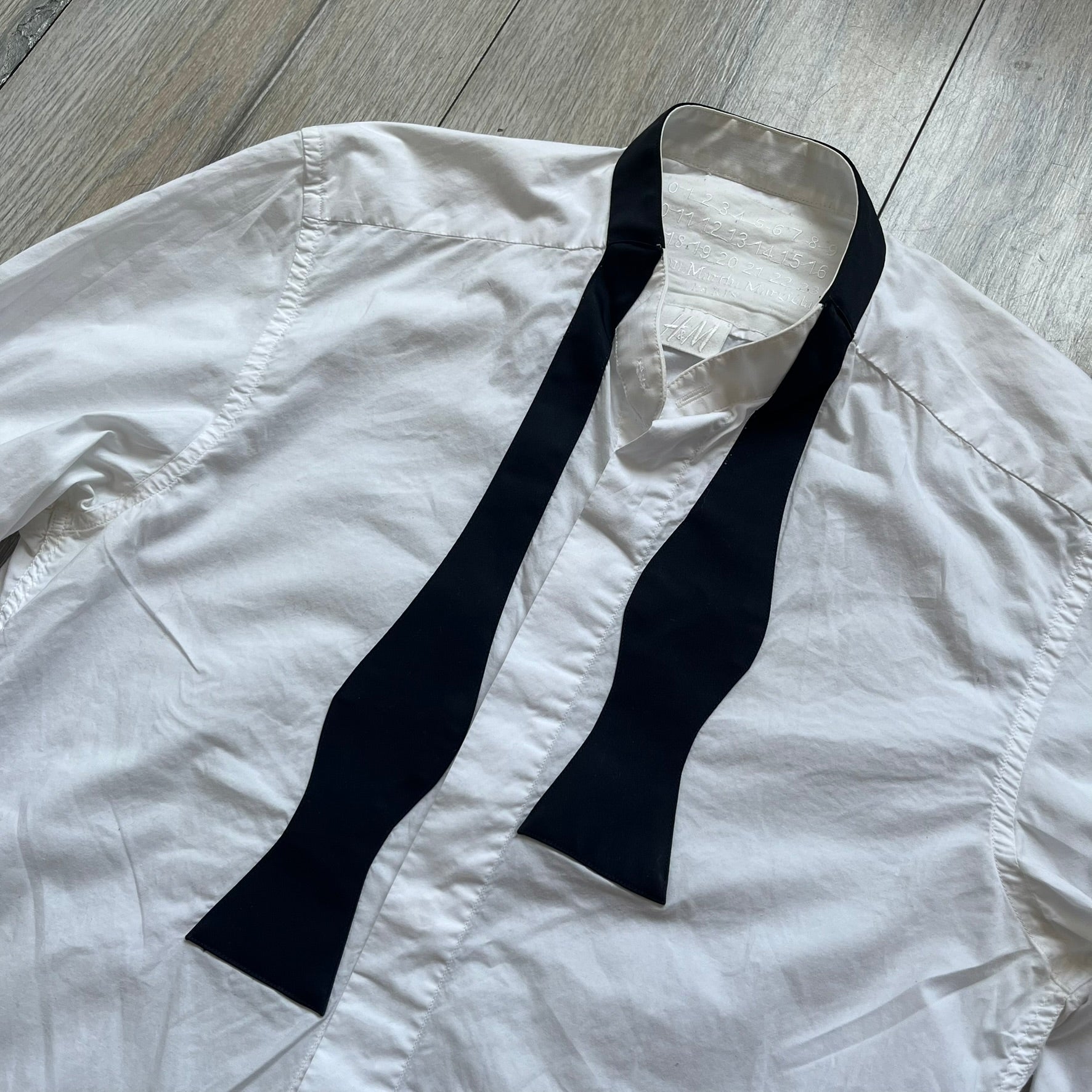 White long-sleeve shirt from the Maison Margiela x H&amp;M Autumn/Winter 2012 collaboration, constructed from 100% cotton. The design features a permanently sewn-in black bow tie positioned loosely at the collar, creating a trompe l’oeil effect that gives the appearance of a casually undone formal look.&nbsp;