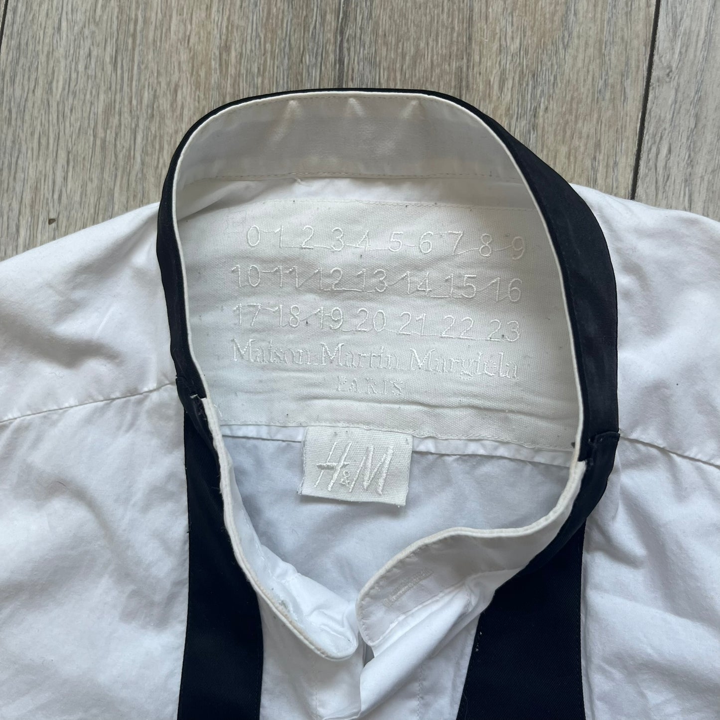 White long-sleeve shirt from the Maison Margiela x H&amp;M Autumn/Winter 2012 collaboration, constructed from 100% cotton. The design features a permanently sewn-in black bow tie positioned loosely at the collar, creating a trompe l’oeil effect that gives the appearance of a casually undone formal look.&nbsp;