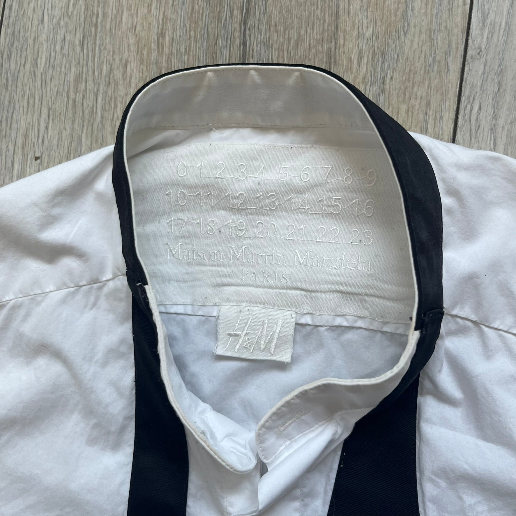 White long-sleeve shirt from the Maison Margiela x H&amp;M Autumn/Winter 2012 collaboration, constructed from 100% cotton. The design features a permanently sewn-in black bow tie positioned loosely at the collar, creating a trompe l’oeil effect that gives the appearance of a casually undone formal look.&nbsp;