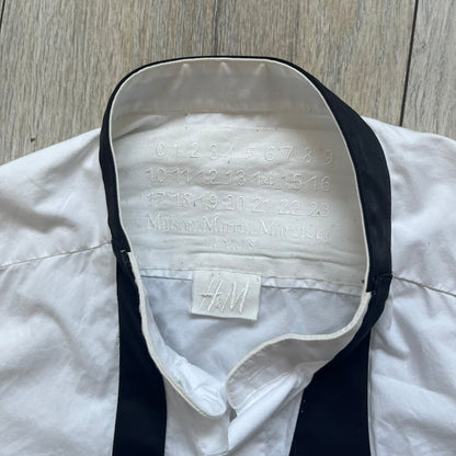 White long-sleeve shirt from the Maison Margiela x H&amp;M Autumn/Winter 2012 collaboration, constructed from 100% cotton. The design features a permanently sewn-in black bow tie positioned loosely at the collar, creating a trompe l’oeil effect that gives the appearance of a casually undone formal look.&nbsp;