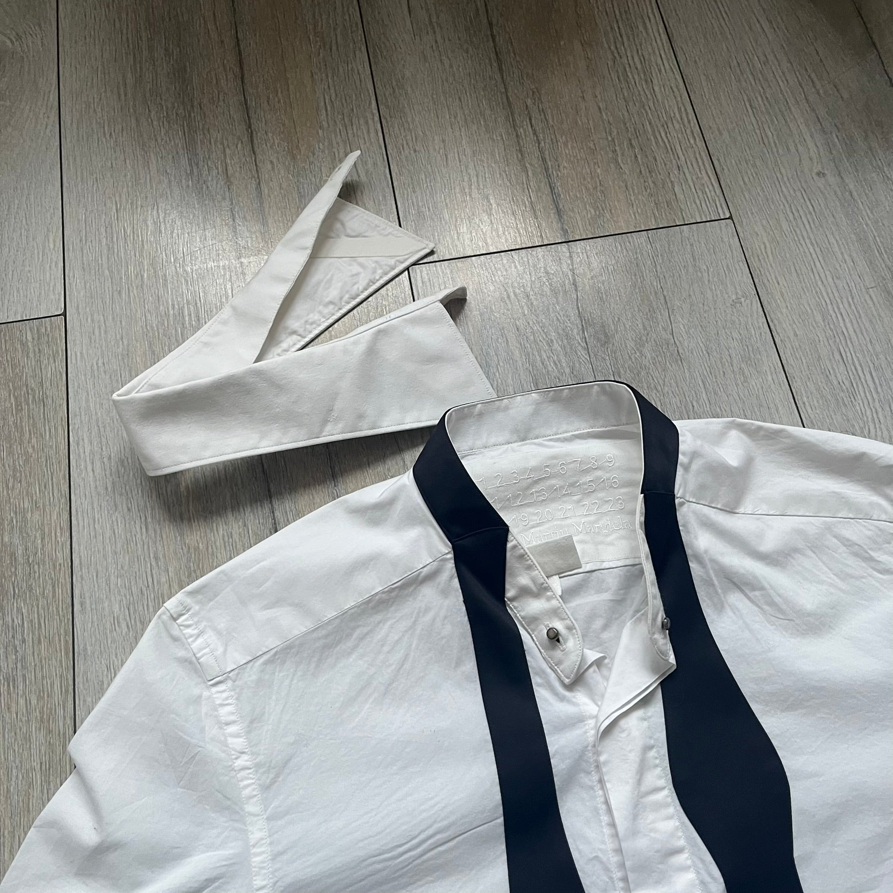 White long-sleeve shirt from the Maison Margiela x H&amp;M Autumn/Winter 2012 collaboration, constructed from 100% cotton. The design features a permanently sewn-in black bow tie positioned loosely at the collar, creating a trompe l’oeil effect that gives the appearance of a casually undone formal look. Additional details include black side panels and underarm stripes for contrast, as well as a detachable collar.