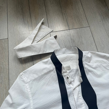 White long-sleeve shirt from the Maison Margiela x H&amp;M Autumn/Winter 2012 collaboration, constructed from 100% cotton. The design features a permanently sewn-in black bow tie positioned loosely at the collar, creating a trompe l’oeil effect that gives the appearance of a casually undone formal look. Additional details include black side panels and underarm stripes for contrast, as well as a detachable collar.