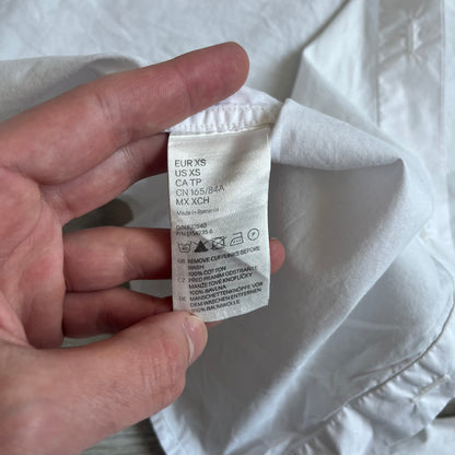 White long-sleeve shirt from the Maison Margiela x H&amp;M Autumn/Winter 2012 collaboration, constructed from 100% cotton. The design features a permanently sewn-in black bow tie positioned loosely at the collar, creating a trompe l’oeil effect that gives the appearance of a casually undone formal look.&nbsp;