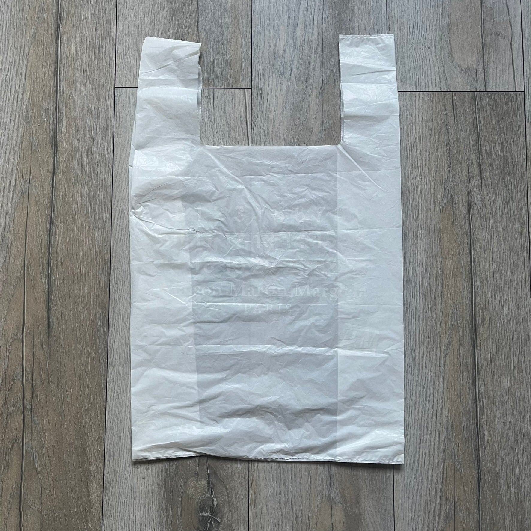 Clear/white plastic bag from the Maison Margiela x H&amp;M collaboration, designed with a cut-off bottom that allows it to be worn as a top or tank. Features big Maison Margiela x H&amp;M logos printed on the front, visible through the semi-transparent material.