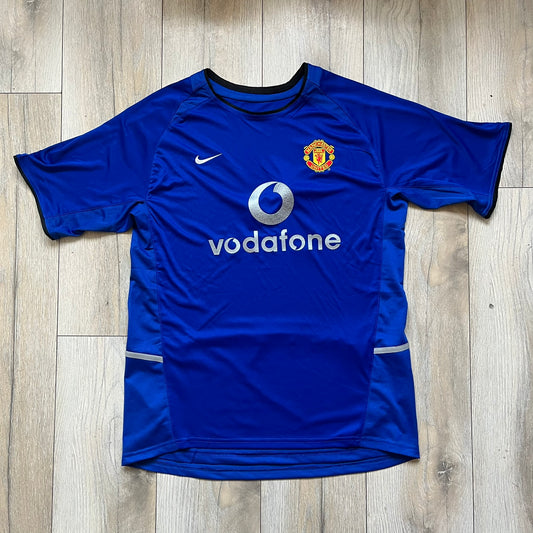 Manchester United 2002/03 Away Retro Blue Tee is a blue short-sleeve shirt from the 2002/03 season, made with Nike Dri-FIT fabric. The design features silver detailing, including a Nike Swoosh on the chest, Manchester United crest on the opposite side, and a central Vodafone sponsor logo. The shirt is made in Morocco.
