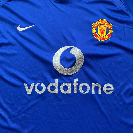 Manchester United 2002/03 Away Retro Blue Tee is a blue short-sleeve shirt from the 2002/03 season, made with Nike Dri-FIT fabric. The design features silver detailing, including a Nike Swoosh on the chest, Manchester United crest on the opposite side, and a central Vodafone sponsor logo. The shirt is made in Morocco.