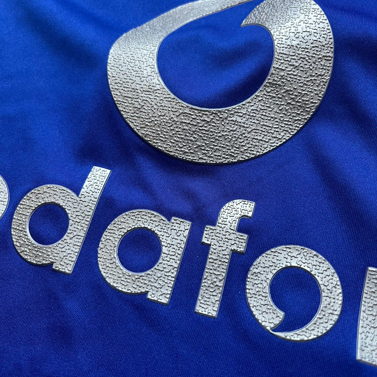 Manchester United 2002/03 Away Retro Blue Tee is a blue short-sleeve shirt from the 2002/03 season, made with Nike Dri-FIT fabric. The design features silver detailing, including a Nike Swoosh on the chest, Manchester United crest on the opposite side, and a central Vodafone sponsor logo. The shirt is made in Morocco.