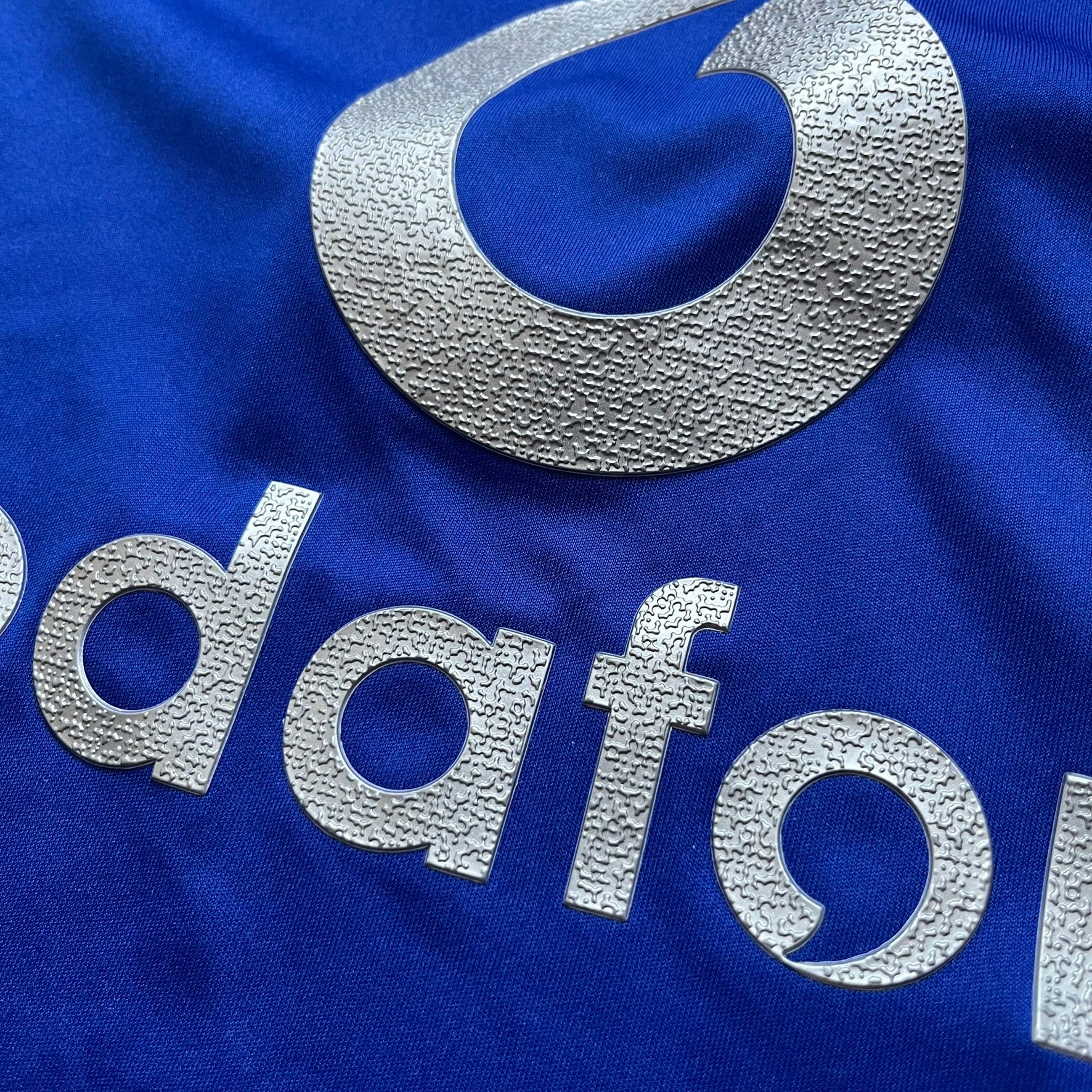 Manchester United 2002/03 Away Retro Blue Tee is a blue short-sleeve shirt from the 2002/03 season, made with Nike Dri-FIT fabric. The design features silver detailing, including a Nike Swoosh on the chest, Manchester United crest on the opposite side, and a central Vodafone sponsor logo. The shirt is made in Morocco.