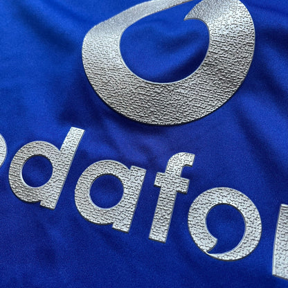Manchester United 2002/03 Away Retro Blue Tee is a blue short-sleeve shirt from the 2002/03 season, made with Nike Dri-FIT fabric. The design features silver detailing, including a Nike Swoosh on the chest, Manchester United crest on the opposite side, and a central Vodafone sponsor logo. The shirt is made in Morocco.