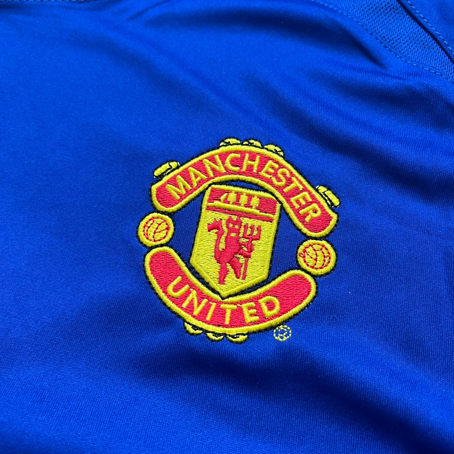 Manchester United 2002/03 Away Retro Blue Tee is a blue short-sleeve shirt from the 2002/03 season, made with Nike Dri-FIT fabric. The design features silver detailing, including a Nike Swoosh on the chest, Manchester United crest on the opposite side, and a central Vodafone sponsor logo. The shirt is made in Morocco.