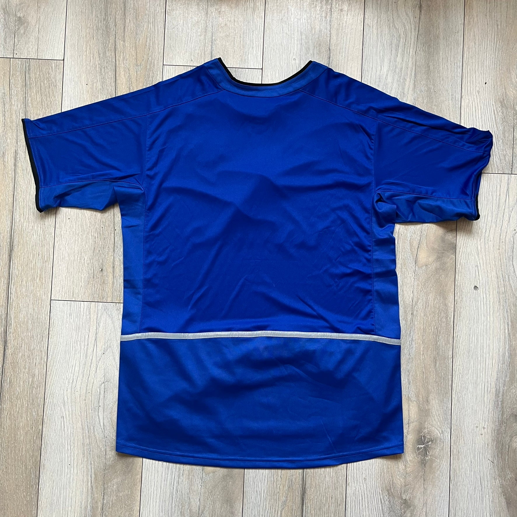Manchester United 2002/03 Away Retro Blue Tee is a blue short-sleeve shirt from the 2002/03 season, made with Nike Dri-FIT fabric. The design features silver detailing, including a Nike Swoosh on the chest, Manchester United crest on the opposite side, and a central Vodafone sponsor logo. The shirt is made in Morocco.