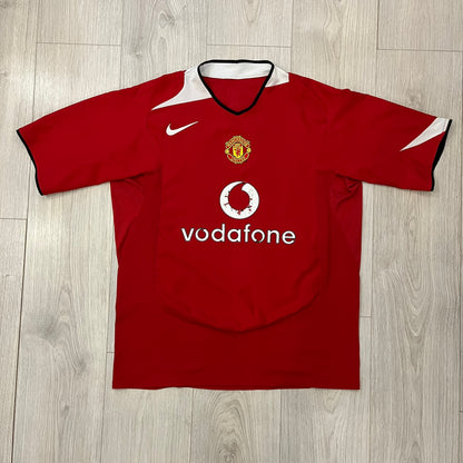 Manchester United 2004/06 Home Football T-Shirt in red, made from Nike Dri-FIT performance fabric. Features the white Vodafone sponsor logo printed across the chest, with an embroidered Manchester United crest above and a white embroidered Nike Swoosh on the right chest. Finished with white contrast detailing around the collar and sleeve hems, matching the official home design used during the 2004–2006 seasons.