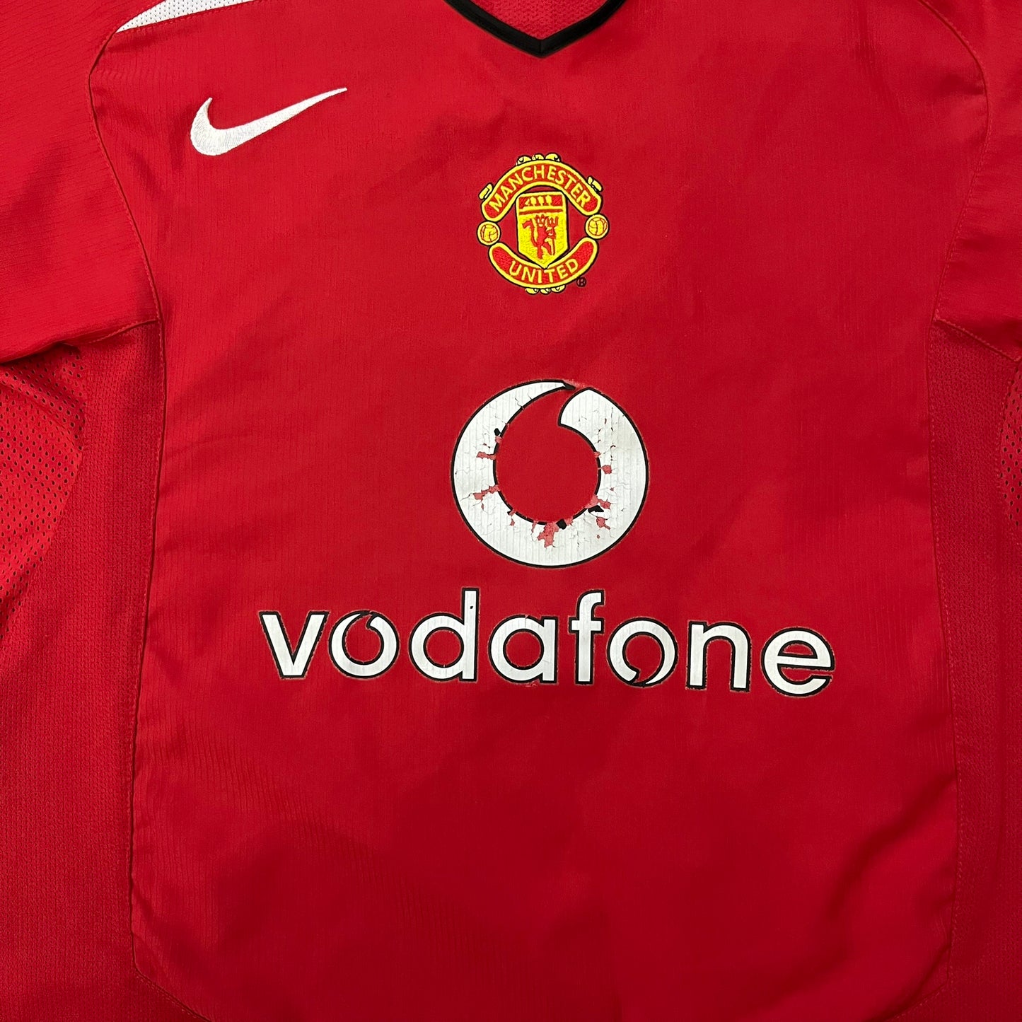 Manchester United 2004/06 Home Football T-Shirt in red, made from Nike Dri-FIT performance fabric. Features the white Vodafone sponsor logo printed across the chest, with an embroidered Manchester United crest above and a white embroidered Nike Swoosh on the right chest. Finished with white contrast detailing around the collar and sleeve hems, matching the official home design used during the 2004–2006 seasons.
