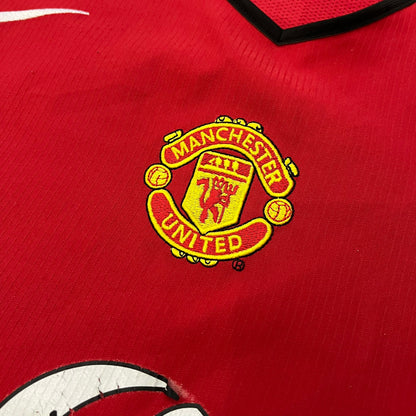 Manchester United 2004/06 Home Football T-Shirt in red, made from Nike Dri-FIT performance fabric. Features the white Vodafone sponsor logo printed across the chest, with an embroidered Manchester United crest above and a white embroidered Nike Swoosh on the right chest. Finished with white contrast detailing around the collar and sleeve hems, matching the official home design used during the 2004–2006 seasons.