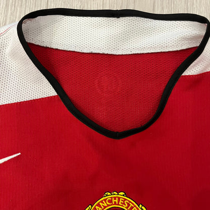 Manchester United 2004/06 Home Football T-Shirt in red, made from Nike Dri-FIT performance fabric. Features the white Vodafone sponsor logo printed across the chest, with an embroidered Manchester United crest above and a white embroidered Nike Swoosh on the right chest. Finished with white contrast detailing around the collar and sleeve hems, matching the official home design used during the 2004–2006 seasons.