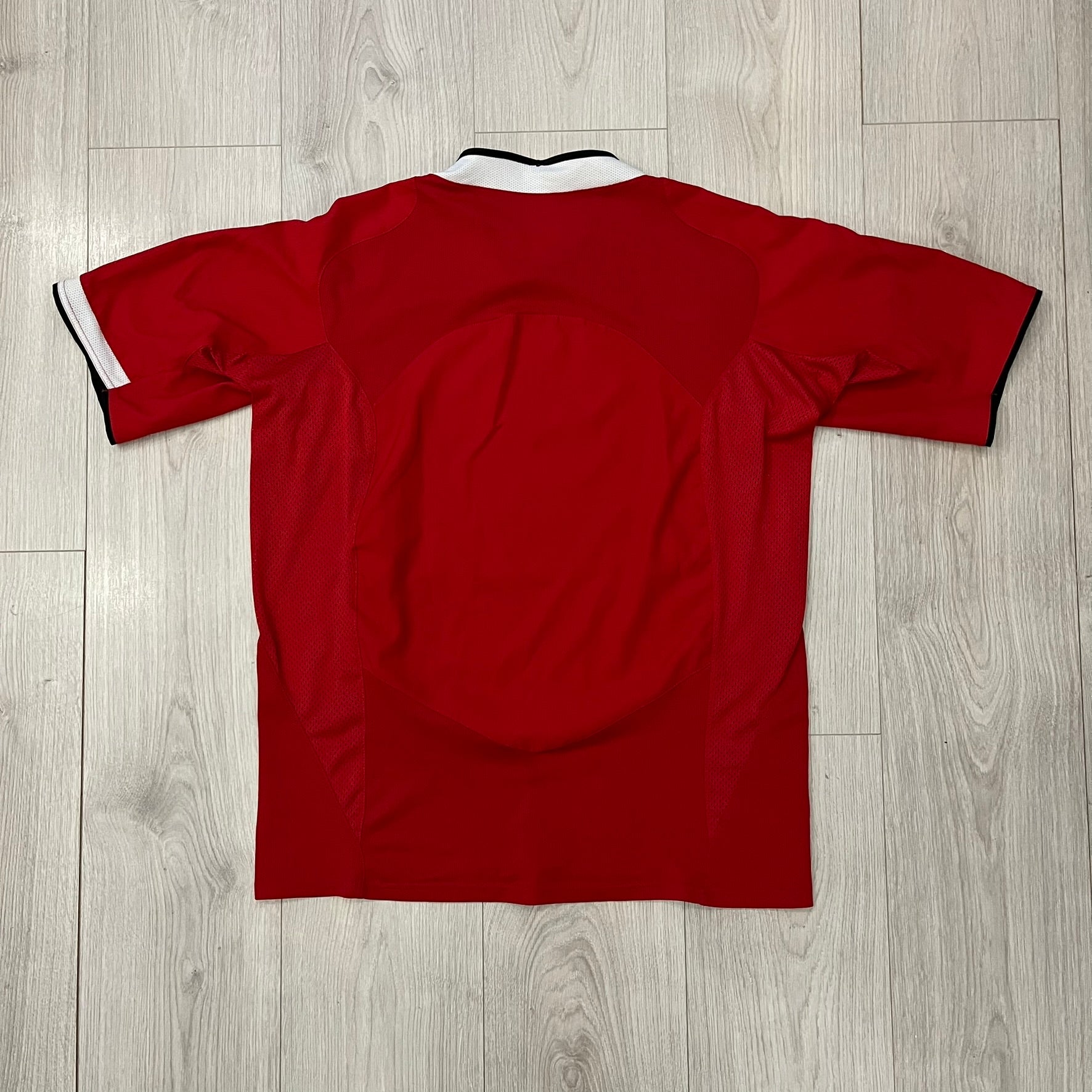 Manchester United 2004/06 Home Football T-Shirt in red, made from Nike Dri-FIT performance fabric. Features the white Vodafone sponsor logo printed across the chest, with an embroidered Manchester United crest above and a white embroidered Nike Swoosh on the right chest. Finished with white contrast detailing around the collar and sleeve hems, matching the official home design used during the 2004–2006 seasons.