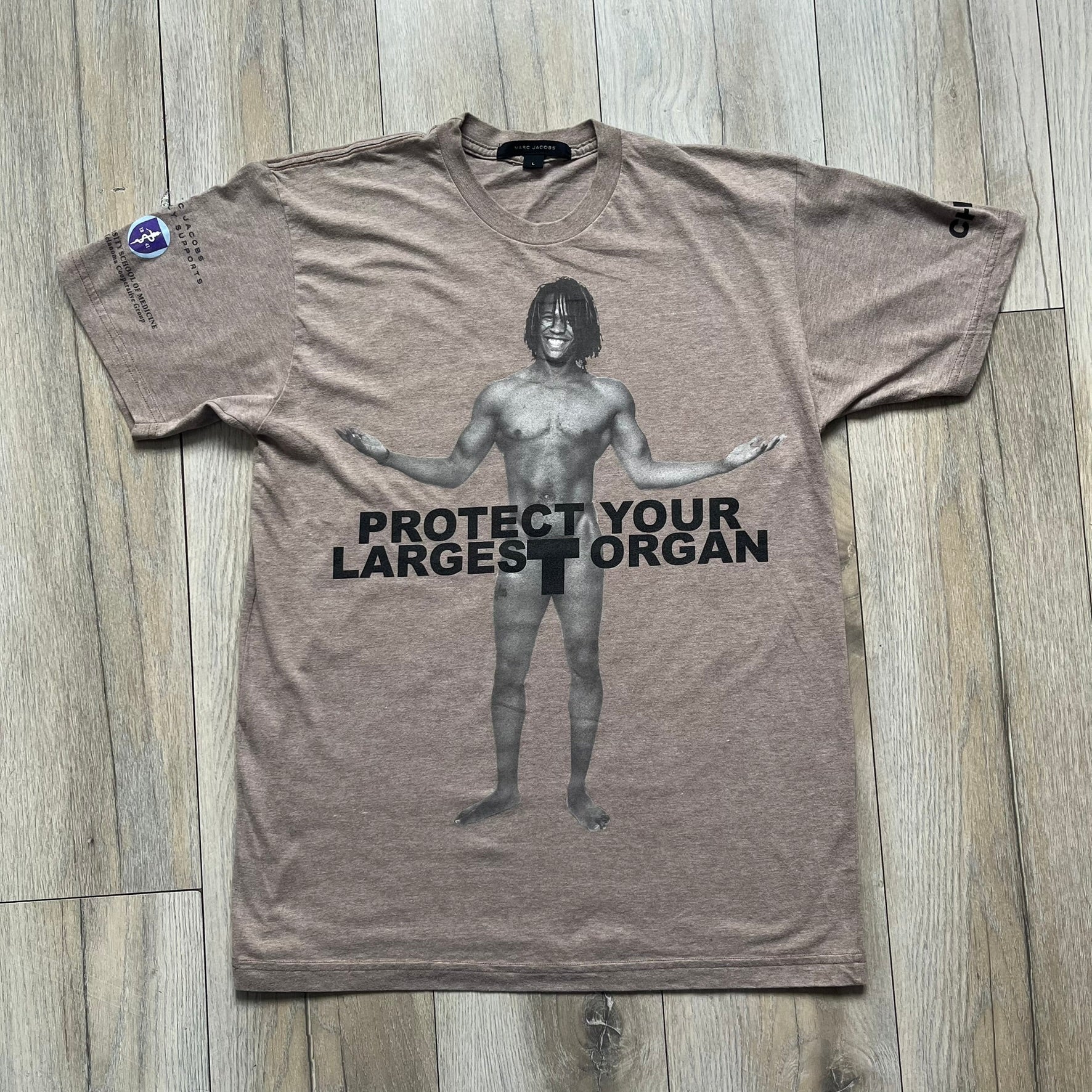 Marc Jacobs 2010s “Protect Your Largest Organ” Chico Clark T-shirt is a light brown awareness campaign tee created to support skin cancer prevention initiatives. The front features an artistic illustration of Chico Clark with accompanying “Protect Your Largest Organ” text. The sleeves include printed details referencing the collaboration and support for NYU School of Medicine. The back displays a large “Thank You” message with credits to contributors involved in the campaign.