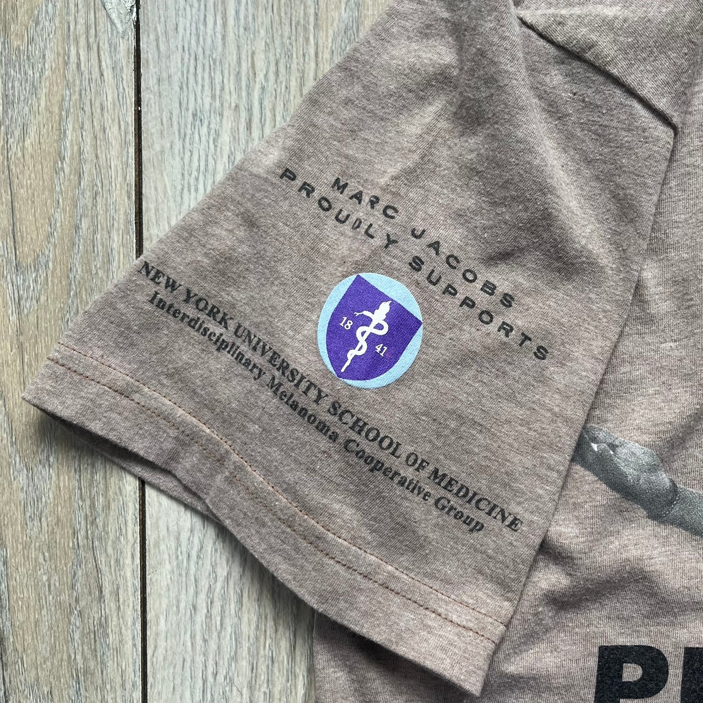 Marc Jacobs 2010s “Protect Your Largest Organ” Chico Clark T-shirt is a light brown awareness campaign tee created to support skin cancer prevention initiatives. The front features an artistic illustration of Chico Clark with accompanying “Protect Your Largest Organ” text. The sleeves include printed details referencing the collaboration and support for NYU School of Medicine. The back displays a large “Thank You” message with credits to contributors involved in the campaign.